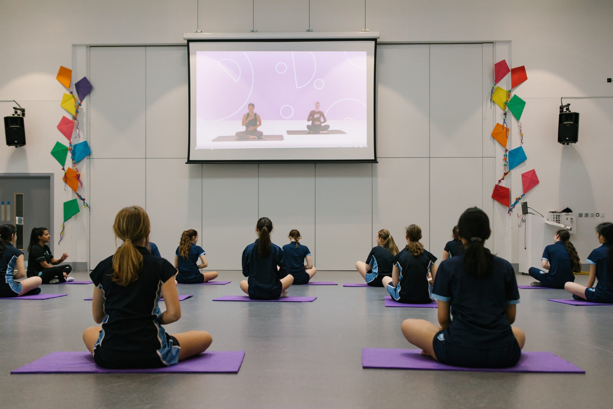 Group of secondary school girls sitting on yoga mats in a PE class, following a Studio You video lesson displayed on a screen.