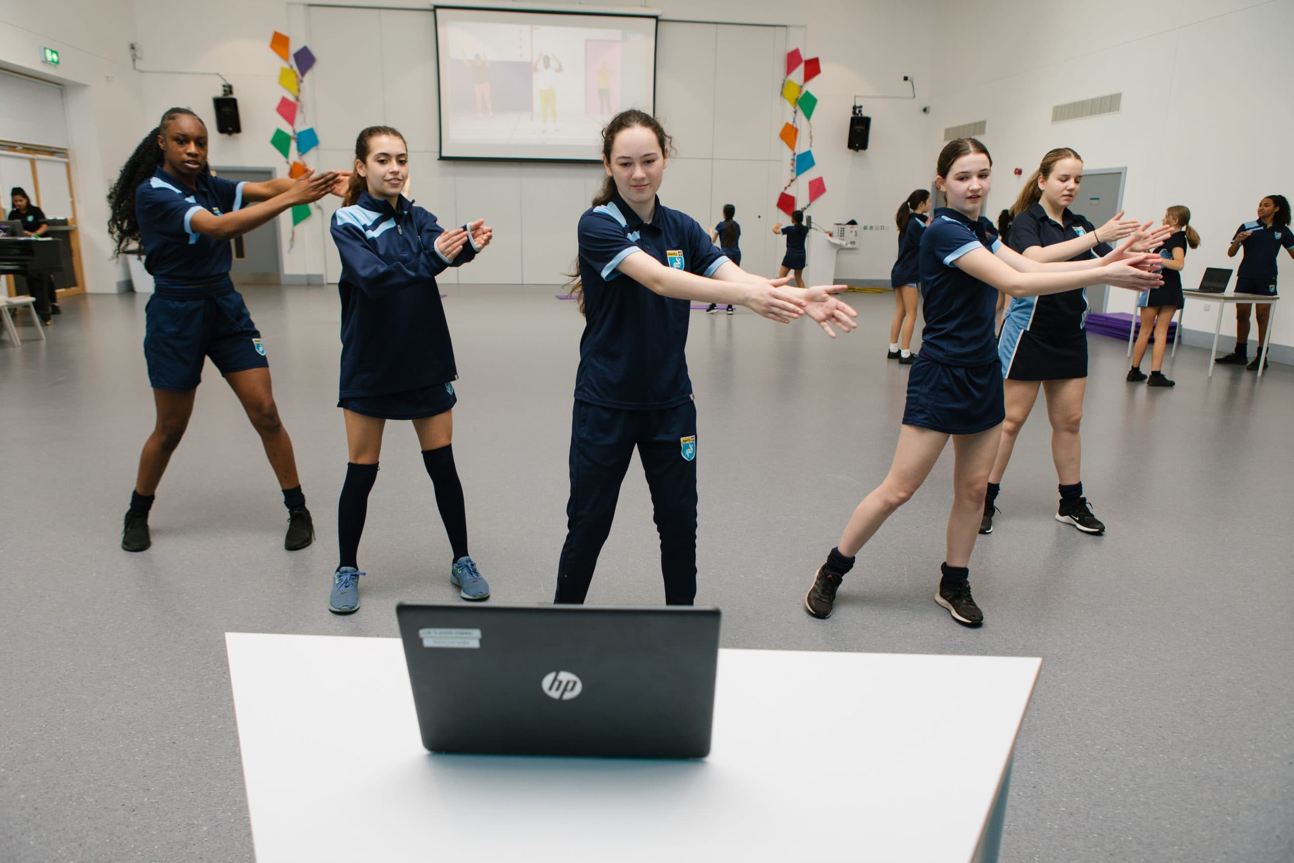 A group of teenage girls dancing while looking at a laptop in a PE class.